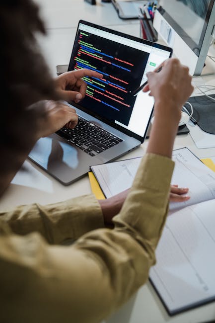 A professional gardener's workspace showing a laptop displaying colorful lines of code, with a person pointing at the screen while seated at a desk. The desk is scattered with papers, a notebook, and 