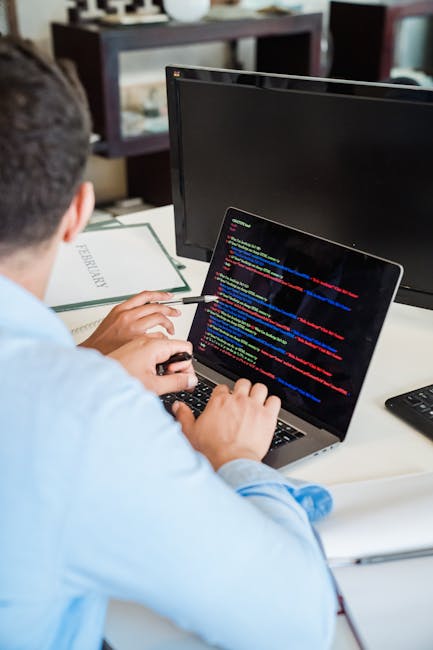 A close-up view of a person's hands using a laptop computer on a dark desk. The screen displays lines of code with syntax highlighting, indicating programming activity. The individual is engaged in ac